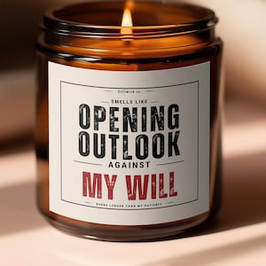 Peut inclure: Une bougie allumée dans un pot en verre brun avec une étiquette blanche. L'étiquette indique "Smells Like Opening Outlook Against My Will". La bougie est posée sur une surface rose.