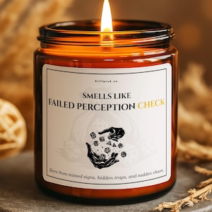 May include: A lit candle in a brown glass jar with a white label. The label reads "SMELLS LIKE FAILED PERCEPTION CHECK" with a graphic of dice and hands. The candle sits on a gray stone surface, with dried plants in the background.