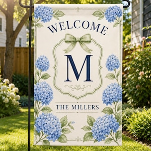 May include: A garden flag featuring the word "WELCOME", a green bow, and the letter "M" within a decorative frame. The flag is adorned with blue hydrangea flowers and the name "THE MILLERS" at the bottom.