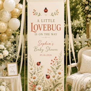 May include: A vertical cream-colored banner featuring the text A LITTLE LOVEBUG IS ON THE WAY, Sophia's Baby Shower, and 05.16.2026. The design includes illustrations of red ladybugs, white daisies, and green foliage, suspended on a gold metal stand.