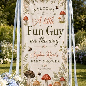 May include: A vertical fabric banner hanging from a white stand with light blue ribbons, featuring the text Welcome to A little Fun Guy on the way, Sophia Ricci's BABY SHOWER, August 12, 2026, decorated with illustrations of mushrooms and bees.