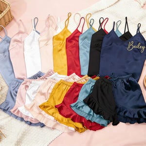 May include: A collection of satin pajama sets in various colors, including blue, pink, white, yellow, red, and black. The sets consist of camisole tops and ruffled shorts. One navy blue set is personalized with the name "Bailey" in gold script.