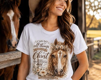 May include: White t-shirt with a horse graphic and the text "I can do all things through Christ who strengthens me" from Philippians 4:13. The shirt features a horse head with a floral crown. The person is wearing a cowboy hat and jeans.