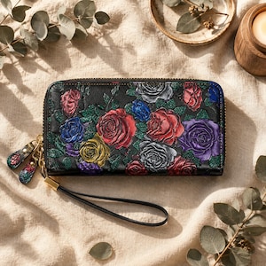 May include: A rectangular black wallet featuring an embossed floral pattern with multi-colored roses in shades of red, blue, yellow, and purple. The wallet includes a gold-toned zipper closure, two matching zipper pulls, and a black wrist strap.