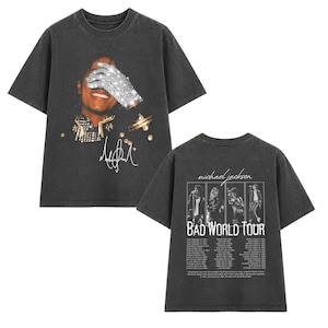 May include: A charcoal gray short-sleeve t-shirt featuring a front graphic of Michael Jackson wearing a sequined glove over his eyes and a signature. The back displays four black and white photos, the text Michael Jackson BAD WORLD TOUR.