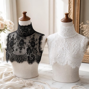 May include: Two lace collar inserts, one black and one white, displayed on mannequin busts. The collars feature intricate floral patterns and high necklines. A pearl necklace and fabric are in the foreground.