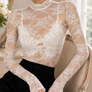 May include: A white long-sleeve mock neck top made of sheer floral lace fabric. The garment features a fitted silhouette with intricate lace patterns throughout the sleeves and bodice, worn over a matching white camisole with thin shoulder straps.
