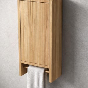 Può includere: Un mobile in legno chiaro montato a parete con un portasciugamani e un asciugamano bianco. Il mobile è di forma rettangolare ed è montato su un muro grigio. Design semplice e moderno.