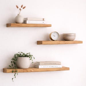 Puede incluir: Tres estantes flotantes de madera con objetos decorativos. El estante superior contiene un pequeño jarrón con flores secas y libros. El estante del medio tiene un reloj dorado y un cuenco. El estante inferior muestra una planta en maceta y libros.