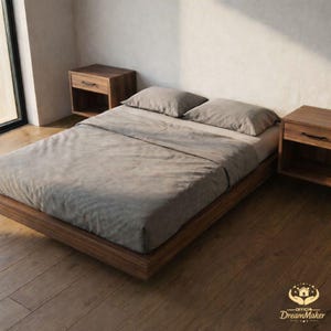 May include: A contemporary bedroom setup with a low wooden bed frame, grey bedding, and two pillows. Two wooden bedside tables with drawers are positioned either side of the bed. The room has wooden flooring and a large window.