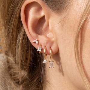 May include: A set of four gold-toned earrings worn in a single ear. The collection includes a small leaf-shaped stud, a small hoop with a flower-shaped charm, a small hoop with a round stone charm, and a small hoop with a rectangular stone charm.