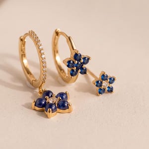 May include: A pair of gold hoop earrings featuring small blue flower-shaped charms. One earring has a flower charm with a central clear stone, while the other features a larger flower charm with blue petals and a clear center stone.
