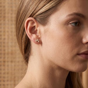 May include: A gold stud earring featuring three small, clear, round-cut stones arranged in a cluster. The earring is positioned on the earlobe, showcasing a metallic gold setting that holds the stones securely in a compact, triangular formation.
