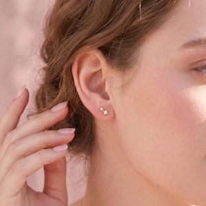 Puede incluir: Primer plano de una oreja adornada con dos pequeños pendientes dorados en forma de estrella. Los pendientes tienen un diseño delicado, con una estrella más pequeña encima de otra más grande, ambas con detalles brillantes.