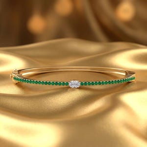 Könnte beinhalten: Ein goldener Armreif mit einer durchgehenden Reihe kleiner, runder, grüner Edelsteine entlang des Bandes, mit einem einzelnen, rechteckigen, klaren Edelstein im Baguetteschliff in der Mitte des Designs und einem sichtbaren Scharnierverschluss.
