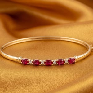 Könnte beinhalten: Ein goldener Armreif mit einer Reihe von vier oval geschliffenen roten Rubinen, die sich mit drei kleinen rund geschliffenen weißen Diamanten abwechseln. Das polierte Goldband verfügt über einen Scharnierverschluss für eine sichere Form.