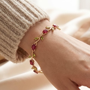 Könnte beinhalten: Ein goldfarbenes Armband mit einem rankenartigen Design, das abwechselnd ovale rote Edelsteine und kleine, marquise-geschliffene grüne Edelsteine aufweist. Die Metallglieder verbinden die Steine in einem flexiblen, organischen Muster.