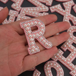 Puede incluir: Letra "R" rosa con adornos de perlas y pedrería. La letra está rodeada por un borde de pedrería y rellena de perlas. Otras letras son visibles en el fondo.