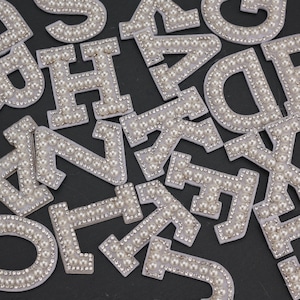 May include: Close-up of various white fabric letters, each embellished with faux pearls and small, clear rhinestones. The letters are scattered across a dark surface, creating a decorative and personalized design element. Ideal for crafting or adding a touch of elegance.