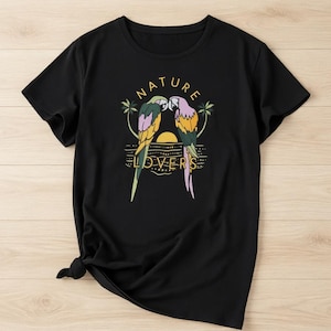 May include: Black t-shirt featuring a colorful graphic of two parrots kissing, with the words "NATURE LOVERS" in a yellow font. The design includes a sunset and palm trees, creating a tropical theme.