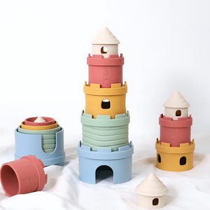 May include: A set of stackable castle-themed building blocks in muted colors including blue, sage green, mustard yellow, terracotta, and cream. The pieces feature cut-out archways, crenelated edges, and embossed numbers, arranged in towers.