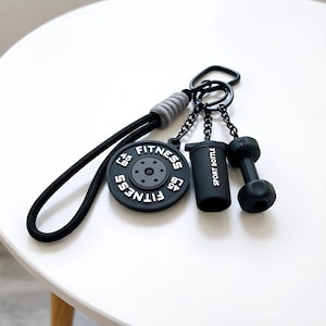 May include: Black fitness-themed keyring with a weight plate, dumbbell, and sport bottle charms. The weight plate is labelled "Fitness" and "45 LB". The keyring has a black cord and a grey accent.