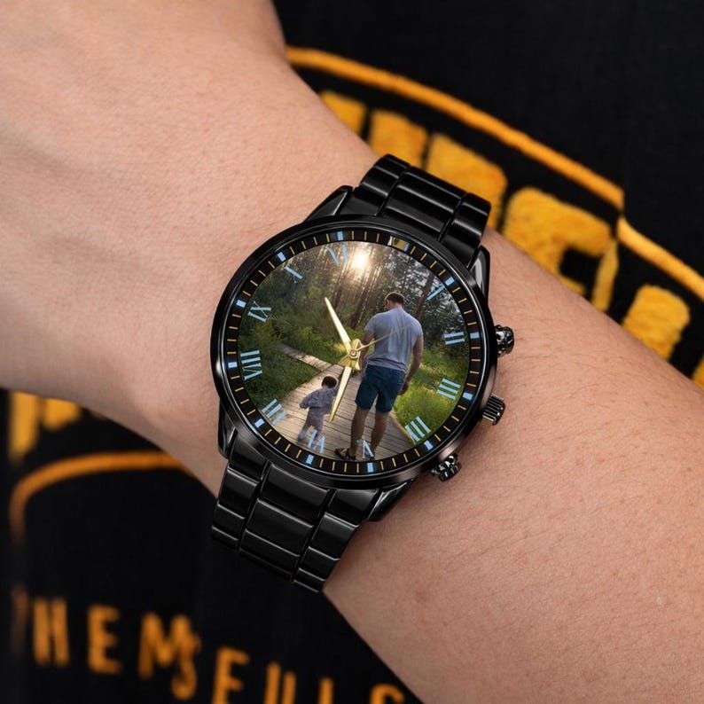 May include: Black wristwatch with a round face featuring a photograph of a person and child walking on a path. The watch has Roman numeral hour markers and gold-colored hands. The watchband is made of black metal links.
