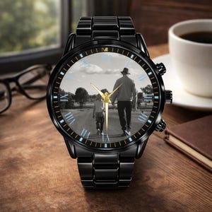 May include: Black wristwatch with a black band and a round face. The watch face features a black and white image of a person walking with a child. Gold-colored hands and blue Roman numerals complete the design.