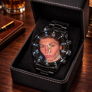 Puede incluir: Reloj de pulsera negro con una correa de acero inoxidable, presentado en una caja negra. La esfera del reloj presenta un retrato con n&uacute;meros romanos y agujas doradas. Un accesorio &uacute;nico.