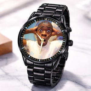 Puede incluir: Reloj de pulsera negro con una correa de acero inoxidable. La esfera del reloj presenta una foto de una persona con expresi&oacute;n de sorpresa, con las manos en la cabeza. El reloj tiene agujas doradas y n&uacute;meros romanos.
