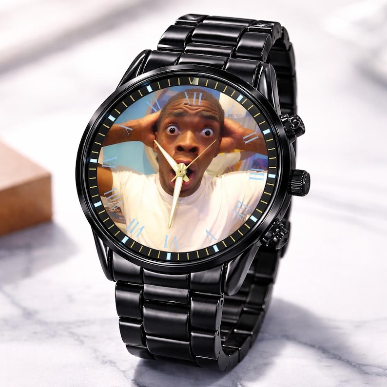Puede incluir: Reloj de pulsera negro con una correa de acero inoxidable. La esfera del reloj presenta una foto de una persona con expresi&oacute;n de sorpresa, con las manos en la cabeza. El reloj tiene agujas doradas y n&uacute;meros romanos.