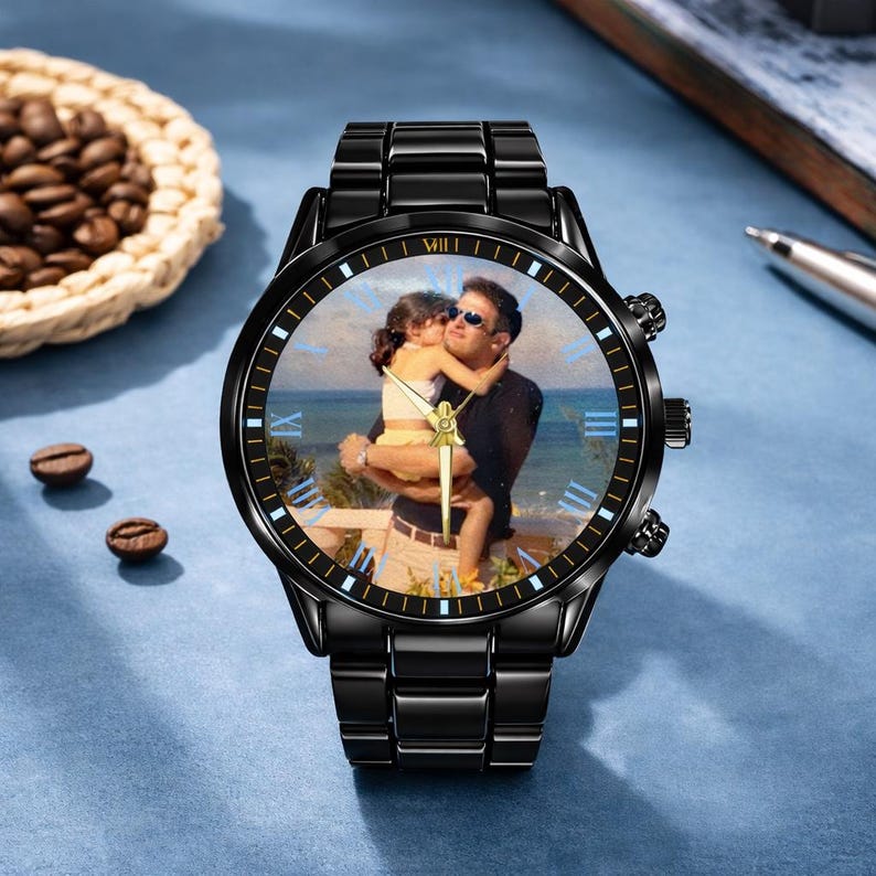 May include: Black wristwatch with a photo of a person holding a child on the watch face. The watch has a black metal band and a round face with Roman numerals. The watch is on a blue surface.