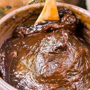 以下が含まれることがあります： 濃い茶色の濃い物質が入った、薄いピンク色のボウルのクローズアップ。木のスプーンがボウルの中にあります。物質は光沢があり、質感のある外観をしています。画像は食品を示唆しています。