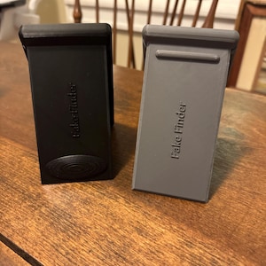 May include: Two 3D-printed phone stands, one black and one gray, are displayed on a wooden surface. The stands have the text "Fake Finder" printed on them. The black stand has a circular design at the bottom.