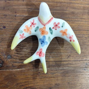 Pode incluir: Estatueta de pássaro de cerâmica branca com asas abertas e cauda bifurcada. O pássaro é decorado com padrões florais coloridos em laranja, rosa e azul. As pontas das asas e a cauda são amarelas. Sobre uma superfície de madeira.