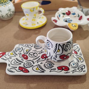 Peut inclure: Une tasse à expresso en céramique blanche et un plateau assorti avec un motif abstrait noir, rouge et jaune. D'autres pièces en céramique avec des motifs floraux et abstraits sont en arrière-plan.