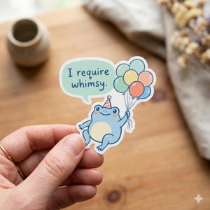 May include: A sticker featuring a light blue frog with a party hat, holding colorful balloons. The frog has a light yellow belly and is in front of a speech bubble that says "I require whimsy."