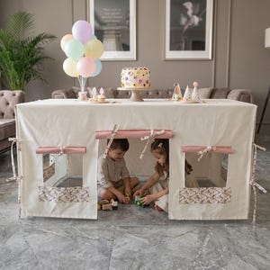 Könnte beinhalten: Ein cremefarbener Stoffüberwurf für einen Spieltisch mit aufrollbaren rosa Fensterrollos und floral gemusterten unteren Fensterpaneelen. Die Konstruktion schafft einen gemütlichen Innenraum zum Spielen mit seitlichen Bindebändern.