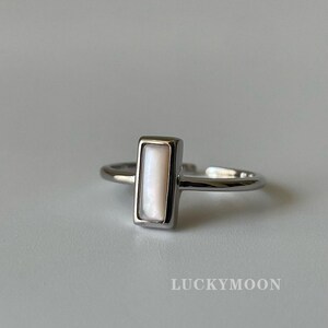 Può includere: Un anello color argento con una montatura rettangolare verticale e un inserto bianco liscio e iridescente. La fascia è aperta sul retro per consentire la regolazione. Il testo LUCKYMOON è stampato nell'angolo in basso a destra.