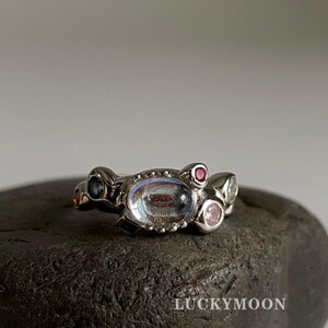 May include: A silver-toned ring featuring an irregular, organic band design. The ring is set with a central oval iridescent stone, flanked by smaller round stones in shades of pink, blue, and clear white. The text LUCKYMOON is visible.