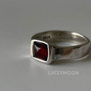 May include: A silver ring featuring a square-cut, deep red gemstone set in a smooth, polished band. The interior of the band is stamped with the text S925. The word LUCKYMOON is visible in the lower right corner of the image frame.