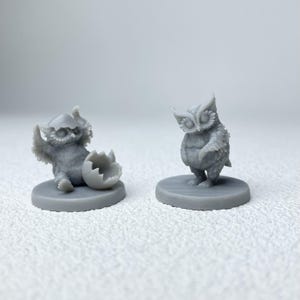 Puede incluir: Dos figuras de b&uacute;hos grises. Un b&uacute;ho emerge de un huevo agrietado, y el otro est&aacute; de pie. Ambas figuras est&aacute;n sobre bases circulares. Los b&uacute;hos tienen texturas de plumas detalladas y ojos grandes.