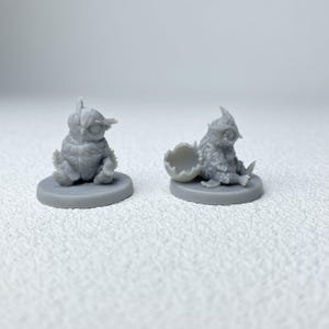 Puede incluir: Dos figuras de b&uacute;ho en miniatura grises. Un b&uacute;ho est&aacute; sentado sobre una base redonda, el otro emerge de una c&aacute;scara de huevo agrietada. Ambas figuras tienen texturas de plumas detalladas y ojos grandes, ideales para juegos de mesa o exhibici&oacute;n.