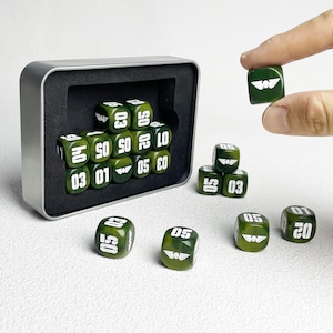 May include: A set of olive green six-sided dice with white numbers and a winged skull emblem. The dice are housed in a rectangular silver metal tin with a black foam insert. Several dice are arranged outside the tin, showing various numerical values.