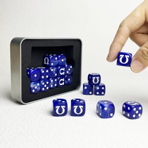 Pode incluir: Um conjunto de dados de seis faces azul translúcido com pontos brancos e o símbolo grego ômega em branco numa das faces. Os dados estão guardados numa caixa metálica retangular prateada com um encaixe de espuma preta.