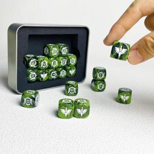 Puede incluir: Un juego de dados de seis caras con efecto mármol verde y negro, con números romanos blancos y un emblema de espada alada. Los dados se guardan en una caja metálica rectangular plateada con espuma negra, con varios dados dispuestos fuera.
