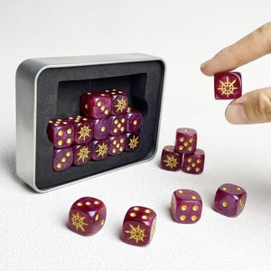 Può includere: Un set di dadi traslucidi viola scuro con puntini dorati e un simbolo a stella a otto punte dorato sulla faccia del sei. I dadi sono contenuti in una scatola di metallo rettangolare argentata con un inserto in schiuma nera.