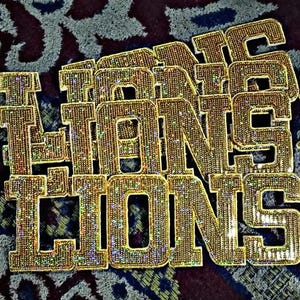 Puede incluir: Colgante dorado con la palabra "LIONS" dispuesta en capas. Las letras están incrustadas con pequeñas piedras brillantes, creando un efecto reluciente. El colgante se encuentra sobre un fondo estampado.