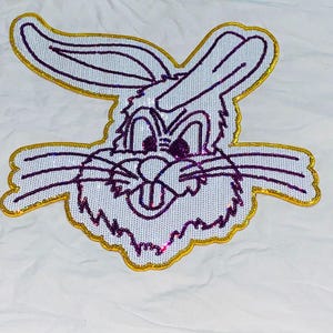 Może przedstawiać: Naszywka z Królikiem Bugsem z białym haftowanym wzorem, fioletowymi detalami i złotą błyszczącą obwódką. Twarz postaci z kreskówki jest wyśrodkowana, z długimi uszami skierowanymi w górę. Odpowiednia do odzieży i akcesoriów.
