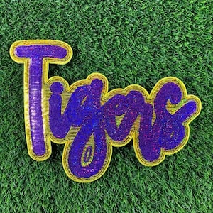 May include: A purple and blue beaded applique with the word "Tigers" in a cursive font. The word is outlined in gold and set against a green grass background. The applique is likely for apparel or accessories.